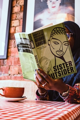 A person is sitting indoors holding a book titled 'Sister Outsider' by Audre Lorde. The cover of the book is green with an abstract illustration of a face. The person's hands are visible, displaying a ring and nail art. The background includes a brick wall and framed artwork. A cup of coffee sits on a saucer on a checkered tablecloth.