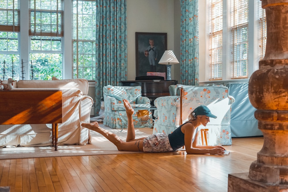 Mujer acostada en el piso marrón cerca del sofá azul y blanco dentro de la habitación blanca con ventanas de vidrio durante el día