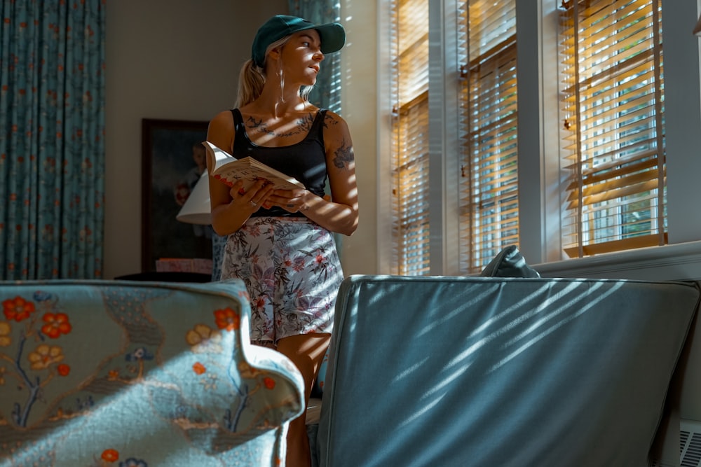 woman holding book while standing beside window blinds