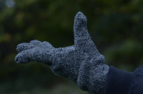 A close-up of a hand wearing a grey, knitted glove with a green, blurred background. The glove is textured and appears to be made of a woolen material, with the hand gesturing or reaching out.