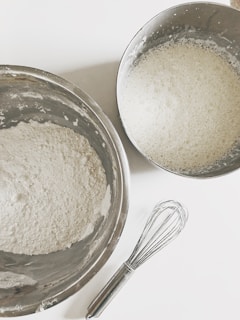 Two stainless steel bowls are placed on a white surface, one containing a white powder that appears to be flour and the other holding a frothy liquid mixture. A metal whisk is positioned nearby, indicating a preparation of ingredients for a baking or cooking activity.