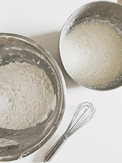 Two stainless steel bowls are placed on a white surface, one containing a white powder that appears to be flour and the other holding a frothy liquid mixture. A metal whisk is positioned nearby, indicating a preparation of ingredients for a baking or cooking activity.
