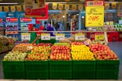 A grocery store display with an assortment of fruits organized in green baskets. The fruits include various types of apples, oranges, and other fresh produce. Price tags and promotional signs are visible above the baskets. A store employee wearing a red cap and green apron is arranging the fruits. In the background, there are shelves with other grocery items and a few shoppers.