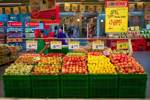 A grocery store display with an assortment of fruits organized in green baskets. The fruits include various types of apples, oranges, and other fresh produce. Price tags and promotional signs are visible above the baskets. A store employee wearing a red cap and green apron is arranging the fruits. In the background, there are shelves with other grocery items and a few shoppers.