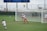 A soccer goalie, wearing a white jersey and red shorts, is in the process of making a save, with a soccer ball in mid-air. A player in a white jersey is approaching from the left while another player stands near the goalpost. The background features a soccer field, a scoreboard, and a large body of water.