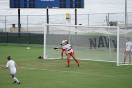 A soccer goalie, wearing a white jersey and red shorts, is in the process of making a save, with a soccer ball in mid-air. A player in a white jersey is approaching from the left while another player stands near the goalpost. The background features a soccer field, a scoreboard, and a large body of water.