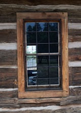 A wooden framed window set into a rustic log cabin wall with visible wood grain and texture. The window is divided into panes by dark grilles and reflects trees and a bit of sky from the outside.