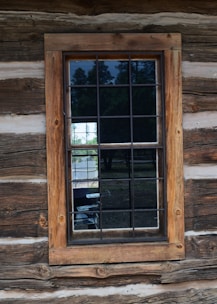 A wooden framed window set into a rustic log cabin wall with visible wood grain and texture. The window is divided into panes by dark grilles and reflects trees and a bit of sky from the outside.