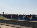 A concrete barrier is in the foreground, and behind it, there's a mural painted on a long wall. The mural includes colorful, abstract shapes and a surreal, humanoid figure with an elongated, stylized face. Above the mural, there's signage for a company, and construction equipment, including a crane, is visible in the distance against a clear sky.