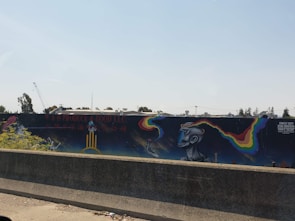 A concrete barrier is in the foreground, and behind it, there's a mural painted on a long wall. The mural includes colorful, abstract shapes and a surreal, humanoid figure with an elongated, stylized face. Above the mural, there's signage for a company, and construction equipment, including a crane, is visible in the distance against a clear sky.