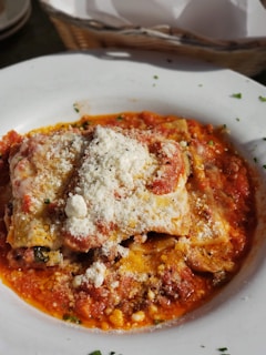 A delicious serving of lasagna topped with a generous amount of grated Parmesan cheese sits on a white plate. The dish is surrounded by a rich, red tomato sauce accented with bits of herbs and spices. The golden-brown layers of pasta and melted cheese create a visually appetizing presentation.