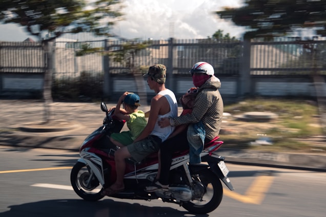 Four people, including a child, ride a motorcycle on a road with blurred background indicating motion. Two adults and a child are seated on the scooter, with one adult wearing a helmet and covering their face. The setting appears urban, with trees and a fence visible in the back.