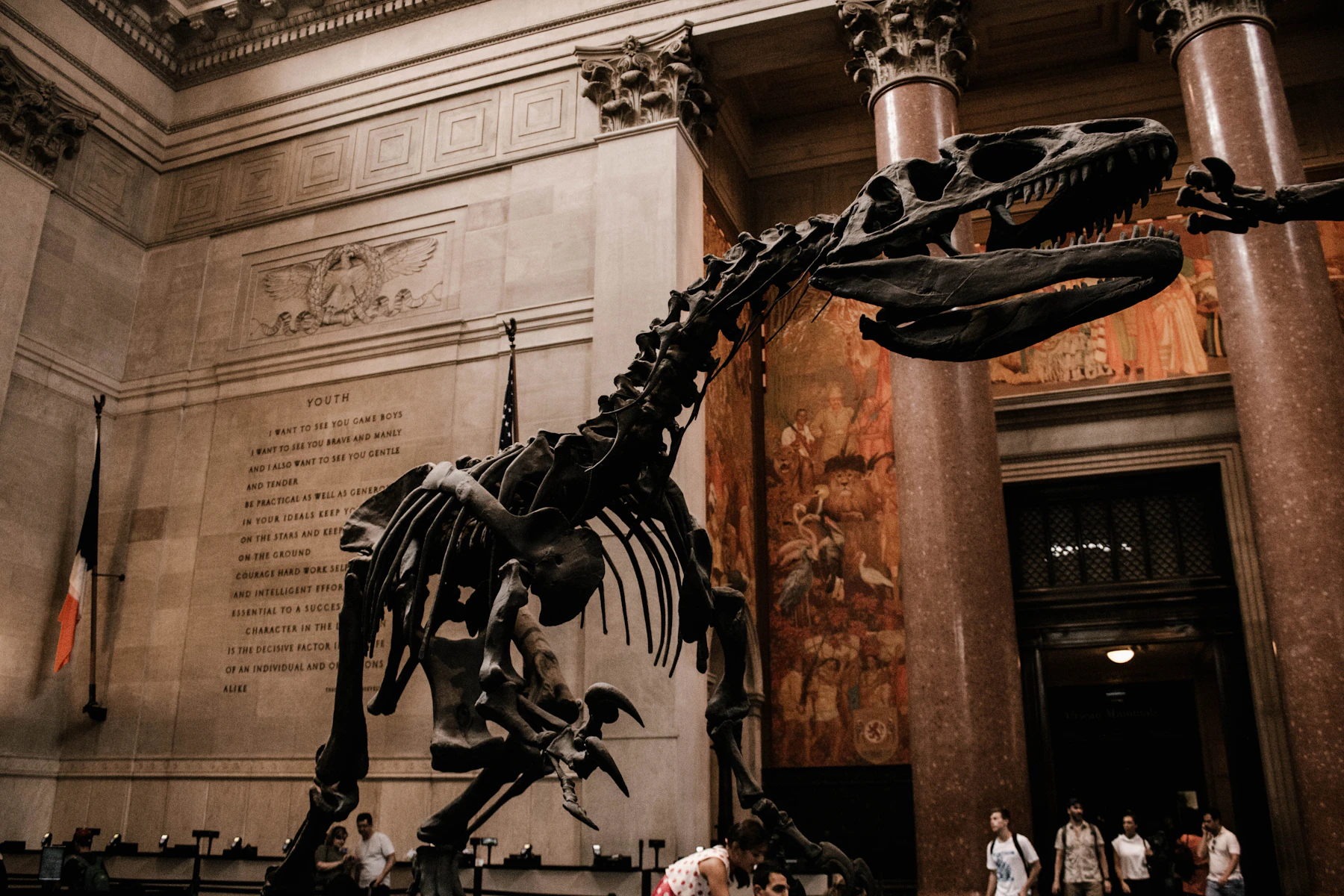 A large dinosaur skeleton mounted in a stone entrance hall between red marble columns, with small groups of visitors at its feet