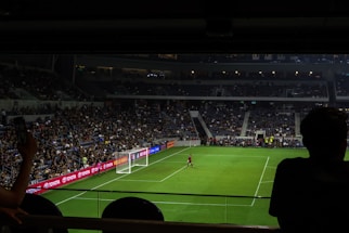 A dimly lit soccer stadium with a large crowd of spectators. A goalkeeper stands near the goalpost while fans are visible in the stands. Advertising boards line the perimeter of the pitch.