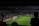 A dimly lit soccer stadium with a large crowd of spectators. A goalkeeper stands near the goalpost while fans are visible in the stands. Advertising boards line the perimeter of the pitch.