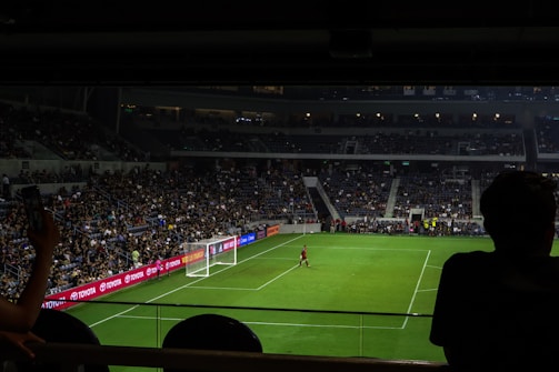 A dimly lit soccer stadium with a large crowd of spectators. A goalkeeper stands near the goalpost while fans are visible in the stands. Advertising boards line the perimeter of the pitch.