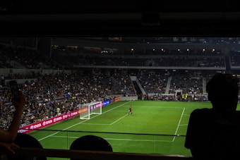 A dimly lit soccer stadium with a large crowd of spectators. A goalkeeper stands near the goalpost while fans are visible in the stands. Advertising boards line the perimeter of the pitch.