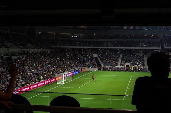 A dimly lit soccer stadium with a large crowd of spectators. A goalkeeper stands near the goalpost while fans are visible in the stands. Advertising boards line the perimeter of the pitch.