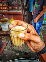 A hand is holding a small glass filled with milky tea. In the background, there are some metal and plastic containers and jars on a wooden surface, and two people are standing nearby. The scene appears to be from a street-side tea stall.