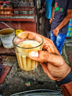 A hand is holding a small glass filled with milky tea. In the background, there are some metal and plastic containers and jars on a wooden surface, and two people are standing nearby. The scene appears to be from a street-side tea stall.