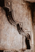 An intricately carved wooden structure with detailed geometric and floral designs, set against a textured wooden background. The craftsmanship suggests a traditional or historical architectural element.