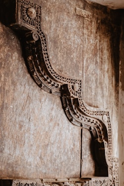 An intricately carved wooden structure with detailed geometric and floral designs, set against a textured wooden background. The craftsmanship suggests a traditional or historical architectural element.