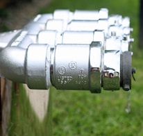 A series of shiny metallic components featuring detailed engravings and labels, indicating technical specifications. The objects appear to be connected in a line, focused in the foreground with a blurred natural background, suggesting they are outdoors.