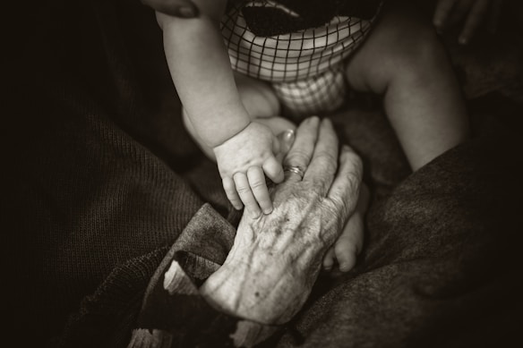 A sepia-toned photograph captures the contrast between a baby's smooth hand resting on an elderly person's wrinkled hand, conveying a sense of connection and generational bond. The baby's checkered outfit adds a subtle pattern, while the elder's hand displays a ring, possibly signifying marriage or tradition.