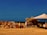 A rustic outdoor market stall features a variety of terracotta pots arranged neatly on the ground. The setup includes a makeshift structure with a tarp providing shade. The background shows a vast expanse of clear sky and barren land, conveying a sense of openness and simplicity.