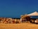 A rustic outdoor market stall features a variety of terracotta pots arranged neatly on the ground. The setup includes a makeshift structure with a tarp providing shade. The background shows a vast expanse of clear sky and barren land, conveying a sense of openness and simplicity.