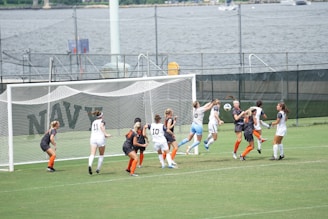 A women's soccer match is taking place near a waterfront. Players from two teams, one in white and the other in dark uniforms with orange socks, are clustered around the goalpost. They seem to be competing for a ball in mid-air, with intense focus on their faces. The background features a fence, water, and distant structures.