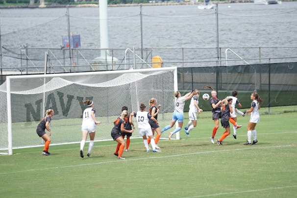A women's soccer match is taking place near a waterfront. Players from two teams, one in white and the other in dark uniforms with orange socks, are clustered around the goalpost. They seem to be competing for a ball in mid-air, with intense focus on their faces. The background features a fence, water, and distant structures.