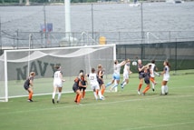 A women's soccer match is taking place near a waterfront. Players from two teams, one in white and the other in dark uniforms with orange socks, are clustered around the goalpost. They seem to be competing for a ball in mid-air, with intense focus on their faces. The background features a fence, water, and distant structures.