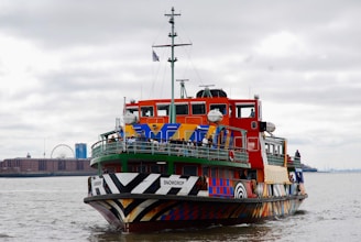 A brightly colored ferry with a unique and artistic design sails on a river. It features a mix of vibrant colors and patterns, attracting attention. Several people are visible onboard, enjoying the view. In the background, a cityscape includes a prominent Ferris wheel and various buildings under an overcast sky.