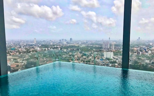 A rooftop infinity pool offers an expansive view of a sprawling cityscape under a partly cloudy sky. Tall buildings and a dense urban landscape spread out in the distance.