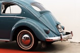 A classic vintage car with a retro design is parked indoors. The vehicle has a teal blue color with a smooth, curved body and distinctive rounded rear end. Chrome accents highlight features like the bumper and hubcap. The car's rear license plate is visible, providing a nostalgic touch.