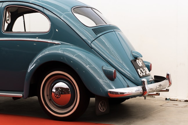 A classic vintage car with a retro design is parked indoors. The vehicle has a teal blue color with a smooth, curved body and distinctive rounded rear end. Chrome accents highlight features like the bumper and hubcap. The car's rear license plate is visible, providing a nostalgic touch.