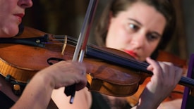 A close-up view of a violinist playing the violin, showing the details of the musician's hands and the strings. The focus is on the elegance and precision of the fingers on the bow and the instrument. A person in the background appears slightly blurred, indicating concentration.