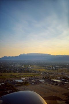 An aerial view of a cityscape with a sprawling urban area in the foreground, transitioning to more green and open spaces further away. In the distance, there is a range of mountains under a clear, slightly hazy sky with a gradient from yellow near the horizon to blue higher up. The image appears to be taken from an airplane, with a part of the engine visible at the lower left corner.