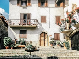 A rustic house with white stucco walls and wooden shutters on the windows. There are two balconies adorned with vibrant red flowers and potted plants. The entrance features an arched wooden door, surrounded by more potted plants and a stone-paved walkway.