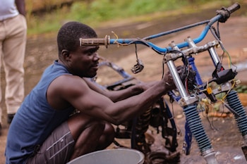 A man wearing a blue sleeveless shirt and shorts is repairing a motorcycle frame. He is focused on adjusting components near the handlebars, with visible wires and parts exposed. The setting appears to be outdoors with a slightly blurred background showing another individual partially, along with some greenery.