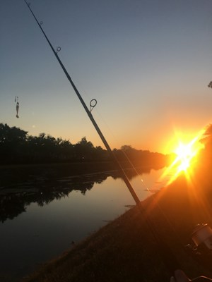 A fishing rod is angled into the frame, positioned next to a tranquil body of water that reflects the warm hues of a setting sun. The sky transitions from orange near the horizon to a deep blue towards the top. Trees line the opposite bank, silhouetted against the bright sunlight.