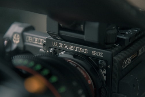 Close-up of a professional video camera with visible branding that reads MONSTRO 8K and RED. The image captures details of the camera's body, lens, and some other components with a slight focus on the text.