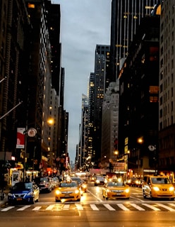 A bustling city street features tall skyscrapers on either side, with numerous vehicles, including iconic yellow taxis, traveling along the road. The scene is taken during twilight, as streetlights and building lights illuminate the urban landscape.