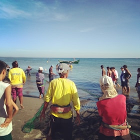 A group of people working together on a beach, pulling nets from the ocean. Some are standing in the water, while others are on the sand. A boat can be seen in the water, indicating a fishing activity. The sky is clear and the water is calm.