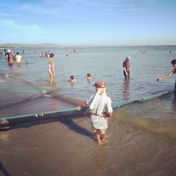 People engage in fishing activities by the shoreline on a sunny day. Some individuals are holding a fishing net, with others, including children, swimming and playing in the shallow water. The scene includes distant hills and a clear blue sky.