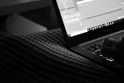 A monochrome image displaying a person typing on a laptop with an illuminated keyboard in a dimly lit room. The screen shows multiple open computer programming or coding interfaces.