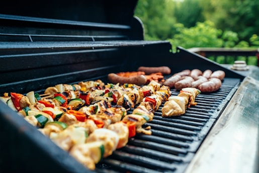 A barbecue grill filled with a variety of foods, including skewered vegetables and meats, as well as sausages. The food is cooking on an open grill with a natural, outdoor setting in the background, featuring lush greenery.