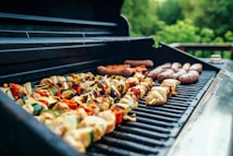 A barbecue grill filled with a variety of foods, including skewered vegetables and meats, as well as sausages. The food is cooking on an open grill with a natural, outdoor setting in the background, featuring lush greenery.