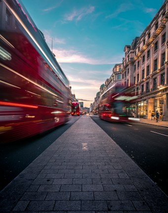 A vibrant urban street scene featuring red double-decker buses in motion. The image captures a bustling atmosphere with blurred motion effect, set against a backdrop of classic architecture and a bright blue sky.